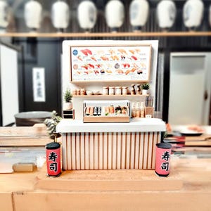May include: Miniature sushi restaurant display featuring a sushi menu, a display case with sushi rolls, and two red and black sushi cans with Japanese text. The counter is made of light wood.