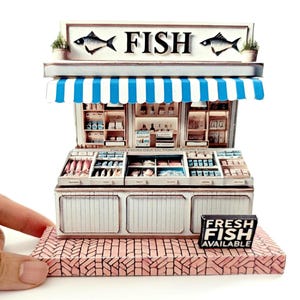 May include: A miniature model of a fish market with a striped awning, a sign that says "FISH", and a sign that says "FRESH FISH AVAILABLE". The model is made of wood and paper and has a detailed interior.