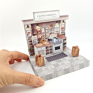 Könnte beinhalten: Ein Miniatur-Papiermodell einer Küche mit einem weißen Herd, Schränken und einem Schild mit der Aufschrift "Ich liebe Kochen". Die Küche ist mit verschiedenen Küchenutensilien und einem kleinen Topf auf dem Herd dekoriert. Das Modell steht auf einer grauen Fliesenfläche.