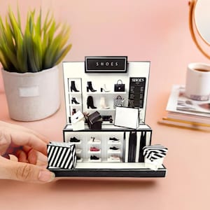 Puede incluir: Miniatura de una zapatería con la palabra "SHOES" en un letrero negro. La exhibición presenta zapatos, bolsos y bolsas de compras diminutos. La tienda es en blanco y negro con un fondo rosa. Una mano sostiene una bolsa de compras a rayas.