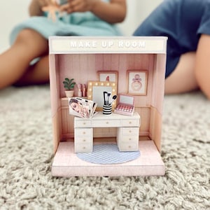 May include: A miniature makeup room playset with a white vanity, a small pink eyeshadow palette, and a black and white striped brush holder. The room has framed artwork and the words "MAKE UP ROOM" on top.