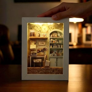 May include: A miniature, illuminated diorama of a cozy room scene, framed in white. The detailed interior features a clock, shelves with miniature items, and a small table. The warm lighting enhances the intricate details of the tiny furnishings.