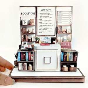 May include: A miniature bookstore diorama with a white counter and shelves filled with books. The sign above the counter reads "BOOKSTORE" and "TOP LIST" with a list of book titles.