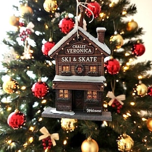 Pode incluir: Um enfeite de árvore de Natal em forma de chalé com o texto "CHALET VERONICA SKI & SKATE". O enfeite castanho tem um telhado com neve, luzes e uma grinalda. É rodeado por bolas vermelhas e douradas.
