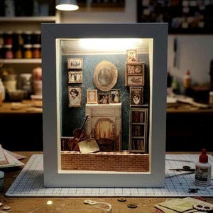 Puede incluir: Una escena de diorama en miniatura dentro de una caja con marco blanco, que presenta una chimenea, fotos enmarcadas, una guitarra y estanterías. La escena está iluminada desde dentro, creando un ambiente cálido. El diorama está sobre una mesa de madera.