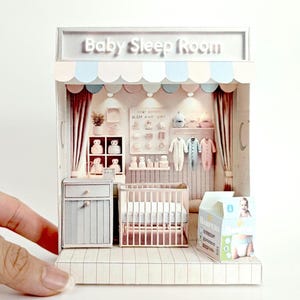 Puede incluir: Una habitación de bebé en miniatura en forma de casa de muñecas. La habitación está decorada en rosa y azul con una cuna, una cómoda y un estante con ropa y juguetes para bebés. El letrero sobre la puerta dice "Baby Sleep Room".