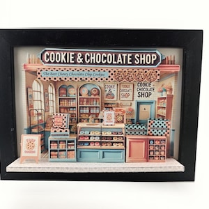 Puede incluir: Diorama en miniatura de una tienda de galletas y chocolate en un marco negro. La tienda cuenta con estanterías repletas de golosinas, una vitrina de donuts y letreros que dicen "Cookie & Chocolate Shop".