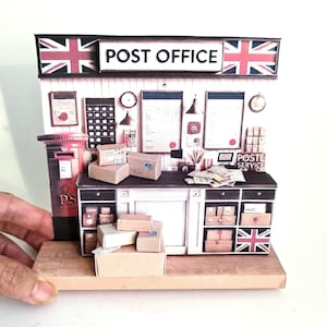 May include: A miniature papercraft model of a British post office with a red post box, a counter with drawers, and shelves stocked with packages. The sign above the post office reads "POST OFFICE" with a Union Jack flag on either side.