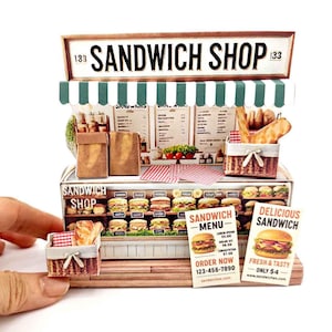 Op de afbeelding: Miniatuurmodel van een broodjeszaak met een groen-wit gestreepte luifel. Het bord leest "SANDWICH SHOP" boven een display van sandwiches, brood en menuborden. Een hand houdt een miniatuurmandje met brood vast.