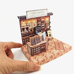 May include: A miniature paper model of a coffee shop with a sign that reads "I love coffee". The shop has a counter with coffee beans and a sign that says "Welcome".
