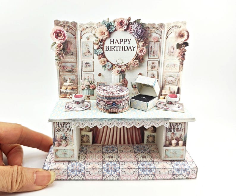 K&ouml;nnte beinhalten: Eine Miniatur-Papierbastel-Szene mit einem wei&szlig;en Tisch, einem floralen Hintergrund und einer Ringbox. Die Szene ist mit Cupcakes, Teetassen und einem Blumenkranz verziert, auf dem "Happy Birthday" steht.