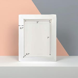 Könnte beinhalten: Ein weißer rechteckiger Bilderrahmen mit einer Öffnung von 12,7 cm x 17,8 cm und einem 1,8 cm breiten Rand. Der Rahmen wird auf einer weißen Oberfläche vor einem rosa und grauen Hintergrund präsentiert.