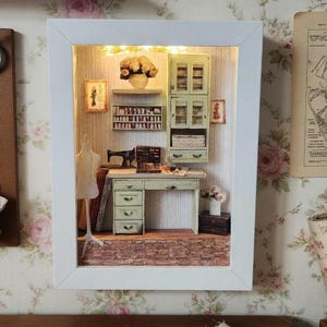 Może przedstawiać: Miniaturowa diorama pracowni krawieckiej w białej ramie. Pokój wyposażony jest w maszynę do szycia, biurko, manekin i półki ze szpulkami nici. W zestawie znajduje się również mała szafka i dzieła sztuki. Tło jest oświetlone małymi światełkami.