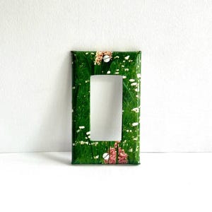 May include: A green light switch cover with a floral design featuring white and pink flowers. The cover has a rectangular shape and a white switch plate.