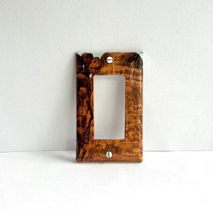 Può includere: Una placca per interruttore con un motivo a venature del legno in tonalità marrone e beige. La placca ha un'unica apertura per l'interruttore e due viti argentate.