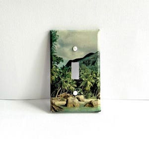 May include: A light switch cover with a tropical beach scene. The image shows a lush green island with palm trees, a rocky beach, and turquoise water.