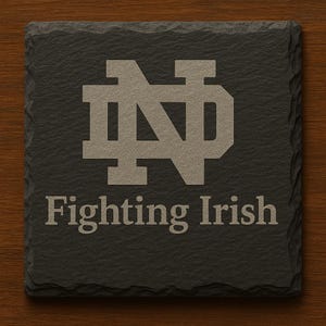 May include: Square, dark gray slate coaster with the words "Fighting Irish" below a tan-colored "ND" logo. The coaster has a rough, textured surface and is set against a wooden background.