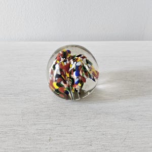 May include: A clear glass paperweight with colorful internal design. The paperweight features a spherical shape with a vibrant arrangement of red, yellow, blue, and white glass elements, creating a decorative and artistic appearance.