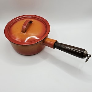 May include: An orange and red enameled cast iron saucepan with a lid and a dark wooden handle. The handle is attached to the side of the pan and has a metal ring at the end. The pan is on a white surface.