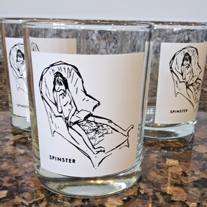 May include: Three clear glass tumblers with a black and white illustration of a person sitting in a chair. The illustration is on a white sticker with the word "SPINSTER" printed below it.