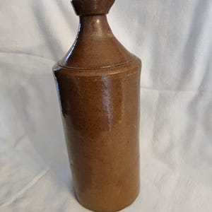 May include: A brown stoneware bottle with a wide mouth and a lid. The bottle is glazed and has a rustic appearance.