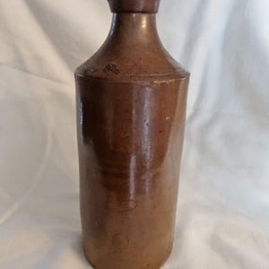 May include: A brown stoneware bottle with a wide mouth and a rounded bottom. The bottle is glazed and has a rustic appearance.