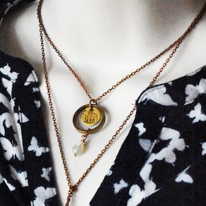 May include: A layered necklace with a bronze chain and pendant. The top layer features a circular pendant with a bear design, and the bottom layer has a small pearl. The necklace is displayed against a black and white butterfly-patterned top.