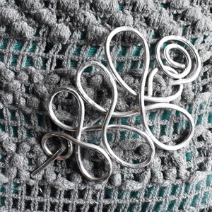 May include: A silver-colored hair barrette with a swirling, abstract design. The barrette is made of metal and is set against a textured, gray crocheted background. The barrette's design includes loops and a pin for securing it in the hair.