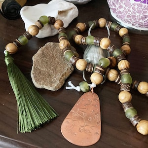 May include: A wooden and green glass beaded necklace with a copper pendant featuring an engraved design. The necklace has a green tassel.