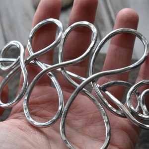 May include: A silver-colored hair accessory with a hammered texture. The metal hair piece has a flowing, abstract design with loops and a spiral. The hair accessory is held in a hand.