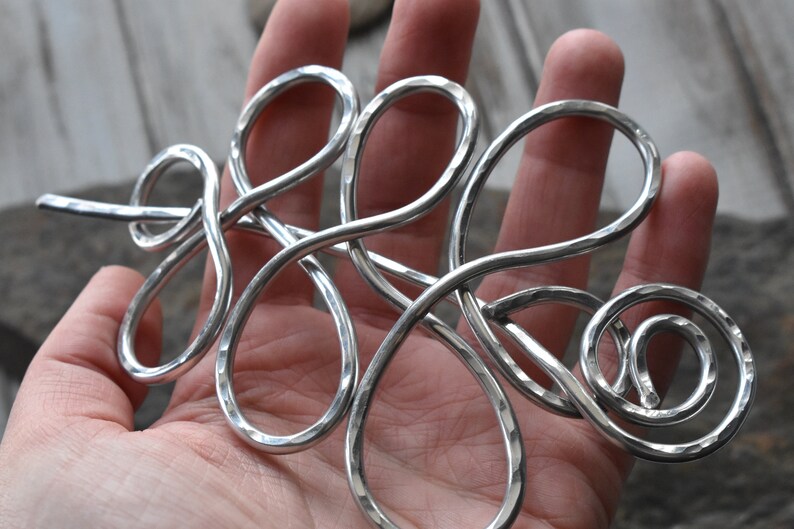 May include: A silver-colored hair accessory with a hammered texture. The metal hair piece has a flowing, abstract design with loops and a spiral. The hair accessory is held in a hand.