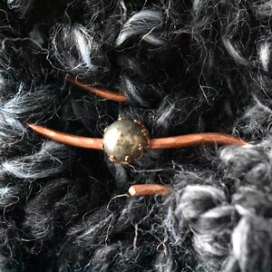May include: Close-up of a hand-knitted textile with a decorative button closure. The yarn is a mix of black, gray, and white. The button is a gold-colored sphere with a copper-colored bar closure. The textile has a textured, fluffy appearance.