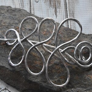 May include: A silver-colored hair barrette with an abstract, flowing design. The barrette is made of curved metal loops and swirls, with a hammered texture. The barrette is displayed on a dark gray stone surface.