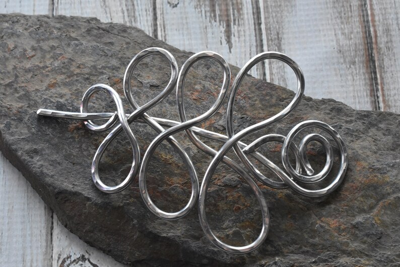 May include: A silver-colored hair barrette with an abstract, flowing design. The barrette is made of curved metal loops and swirls, with a hammered texture. The barrette is displayed on a dark gray stone surface.