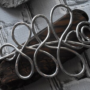 May include: A silver-colored hair barrette with an abstract, flowing design. The barrette is made of a hammered metal and rests on a piece of dark wood. The background is a light gray, textured surface.