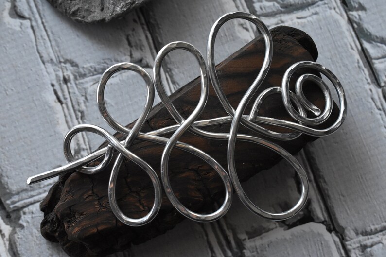 May include: A silver-colored hair barrette with an abstract, flowing design. The barrette is made of a hammered metal and rests on a piece of dark wood. The background is a light gray, textured surface.