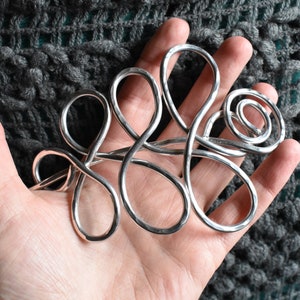 May include: A silver-colored, abstract metal hair accessory with looping and swirling designs. The handcrafted piece is held in a hand, showcasing its intricate form. The background is a textured, gray fabric.