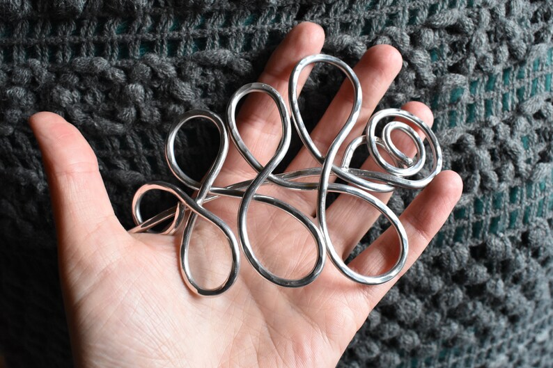 May include: A silver-colored, abstract metal hair accessory with looping and swirling designs. The handcrafted piece is held in a hand, showcasing its intricate form. The background is a textured, gray fabric.