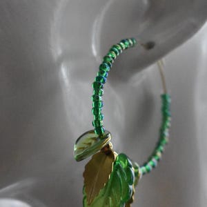 May include: Green and gold leaf-shaped hoop earrings. The earrings are made of small green beads and feature three gold and green leaf charms.