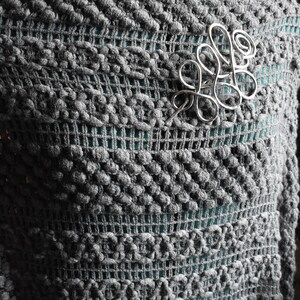 May include: A gray, textured knit shawl with a silver-colored wirework brooch. The shawl has a unique pattern with raised, bobble-like stitches and a mesh-like design. The brooch is an abstract, swirling design.