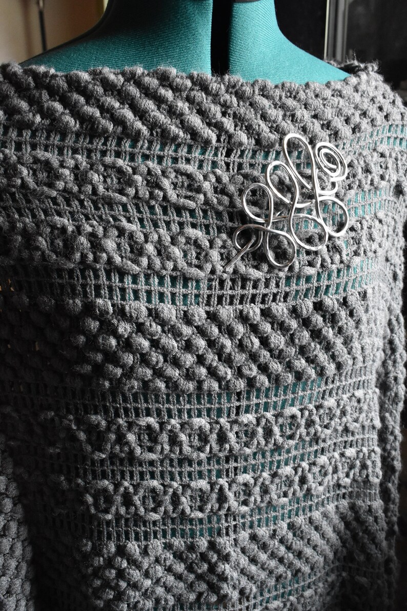 May include: A gray, textured knit shawl with a silver-colored wirework brooch. The shawl has a unique pattern with raised, bobble-like stitches and a mesh-like design. The brooch is an abstract, swirling design.