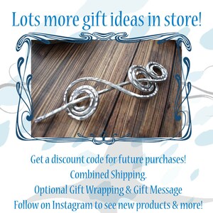 May include: A silver-colored hair stick with a spiral and loop design. The hair accessory is displayed on a wooden surface. The image includes the text "Lots more gift ideas in store!"