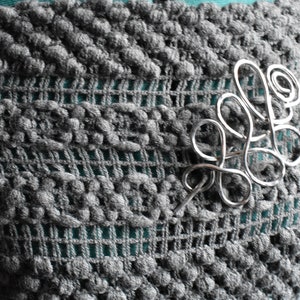 May include: A silver-colored wire brooch in a flowing, abstract design is pinned to a gray, textured fabric. The fabric has a knitted, open weave pattern with a teal-colored underlay visible.