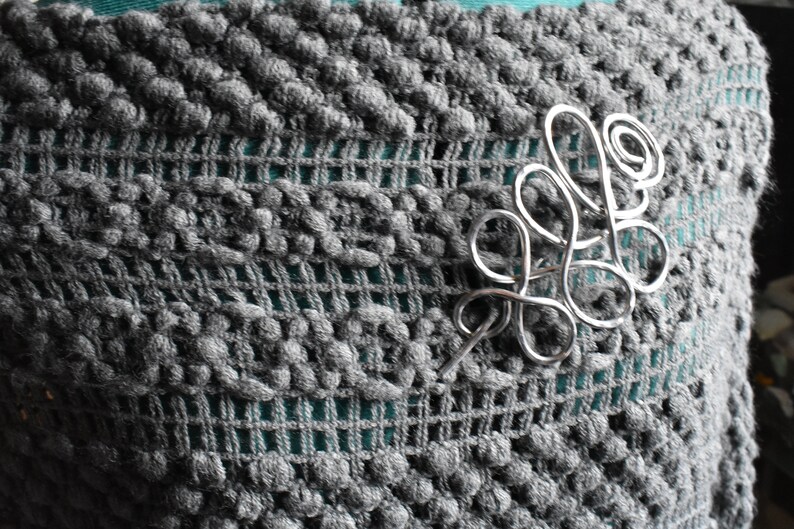 May include: A silver-colored wire brooch in a flowing, abstract design is pinned to a gray, textured fabric. The fabric has a knitted, open weave pattern with a teal-colored underlay visible.