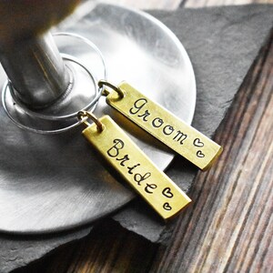 May include: Two gold-colored metal wine glass charms with attached rings. One charm says "Bride" with a heart and the other says "Groom" with two hearts.