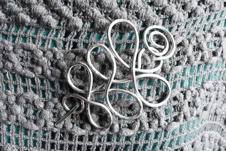 May include: A silver-colored hair barrette with a swirling, abstract design. The barrette is made of metal and is set against a textured, gray crocheted background. The barrette's design includes loops and a pin for securing it in the hair.