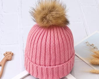 May include: A ribbed knit beanie in a soft pink color, featuring a thick folded cuff at the base and a large, fluffy, light brown faux fur pom-pom attached to the top center of the hat, creating a textured and layered appearance.