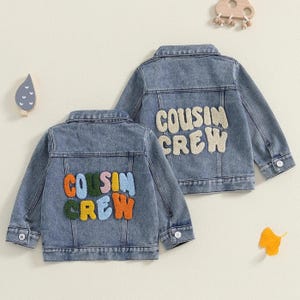 Könnte beinhalten: Zwei hellblaue Jeansjacken mit den Worten "COUSIN CREW" in bunten und cremefarbenen Buchstaben auf dem Rücken gestickt. Die Jacken haben Knopfverschlüsse und Kragen. Die Jacken sind für Kinder.