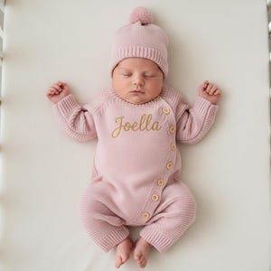 Könnte beinhalten: Ein Baby in einem hellrosa Strick-Strampler und passendem Hut mit Bommel. Der Strampler hat Holzknöpfe und den Namen "Joella" in Goldfaden gestickt. Das Baby liegt auf einer weißen Oberfläche.