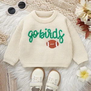 Könnte beinhalten: Cremefarbener Strickpullover mit den grünen Worten "go birds" und einer braunen Football-Grafik. Der Pullover wird mit weißen Turnschuhen und Sonnenbrillen kombiniert, ideal für ein junges Kind.